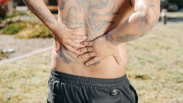 A man with a large tattoo on his back holds his lower back outdoors, showing discomfort.
